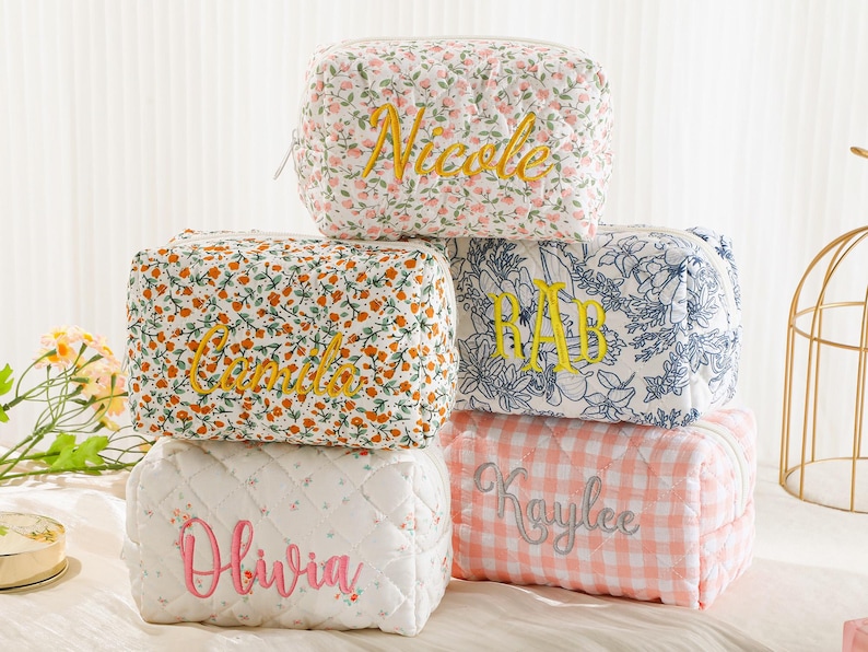 May include: A stack of quilted cosmetic bags with embroidered names. The bags feature floral and gingham patterns in various colors, including white, orange, and pink. The names "Nicole", "Camila", "RAB", "Olivia", and "Kaylee" are embroidered on the bags.