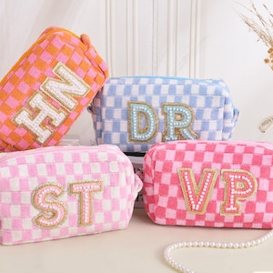 May include: Four checkered cosmetic bags in pink, orange, and blue. Each bag features a different set of initials: HN, DR, ST, and VP, embellished with beads. The bags are rectangular with a zipper closure, perfect for storing makeup or toiletries.