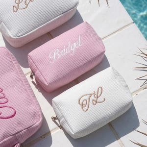 May include: A collection of rectangular waffle-textured pouches in white and soft pink. One pink pouch features the name Bridget embroidered in white script, while a white pouch displays the monogram TL in tan script. Each pouch has a metal zipper.