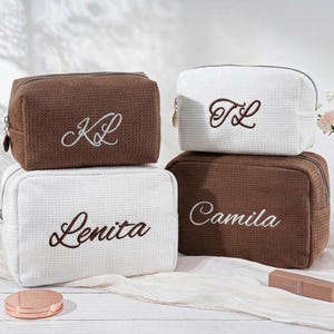 May include: Four rectangular cosmetic bags with a waffle-textured fabric. Two bags are brown and two are white, each featuring embroidered lettering. The brown bags display the initials KL and the name Camila, while the white bags show TL and Lenita.