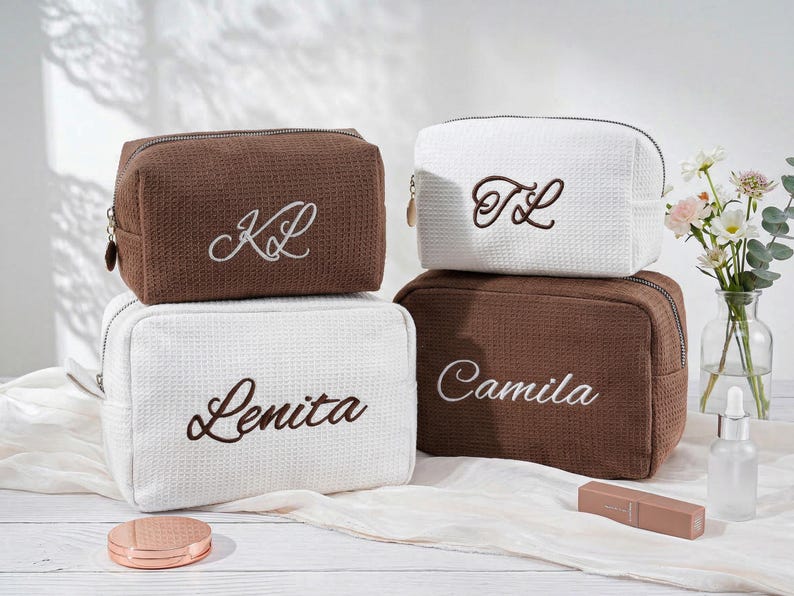 May include: Four rectangular cosmetic bags with a waffle-textured fabric. Two bags are brown and two are white, each featuring embroidered lettering. The brown bags display the initials KL and the name Camila, while the white bags show TL and Lenita.