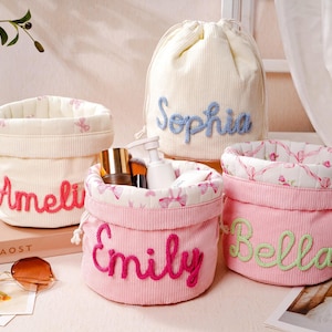 May include: A collection of four fabric storage baskets and one drawstring pouch. The baskets are cream and pink corduroy with embroidered names Ameli, Emily, and Bella. The pouch is cream corduroy with the name Sophia embroidered in blue.