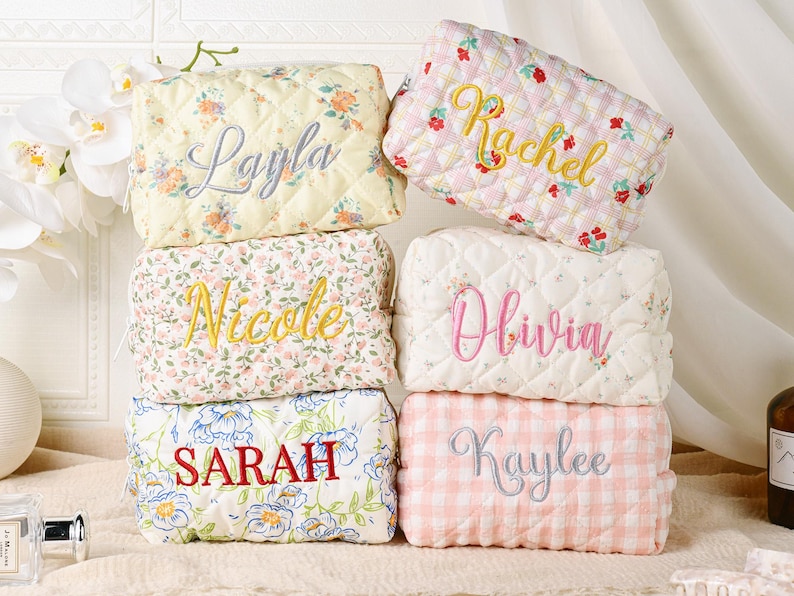 May include: A stack of six quilted cosmetic bags with embroidered names. The bags feature floral patterns in various colors, including yellow, pink, and blue. Each bag has a different name embroidered in a contrasting color.