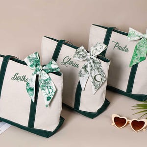 Peut inclure: Trois sacs cabas en toile blanche avec des bordures vertes et des noms personnalis&eacute;s&nbsp;: Bertha, Gloria et Paula. Chaque sac est orn&eacute; d&rsquo;un ruban &agrave; motif vert et blanc nou&eacute; en un n&oelig;ud.