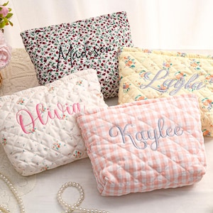 May include: Four quilted cosmetic bags with embroidered names. One bag has a red floral pattern with "Madison" embroidered in black. Another is cream with pink floral and "Olivia" in pink. A yellow floral bag has "Layla" in gray, and a pink gingham bag has "Kaylee" in gray.