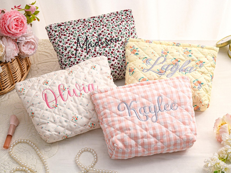 May include: Four quilted cosmetic bags with embroidered names. One bag has a red floral pattern with "Madison" embroidered in black. Another is cream with pink floral and "Olivia" in pink. A yellow floral bag has "Layla" in gray, and a pink gingham bag has "Kaylee" in gray.