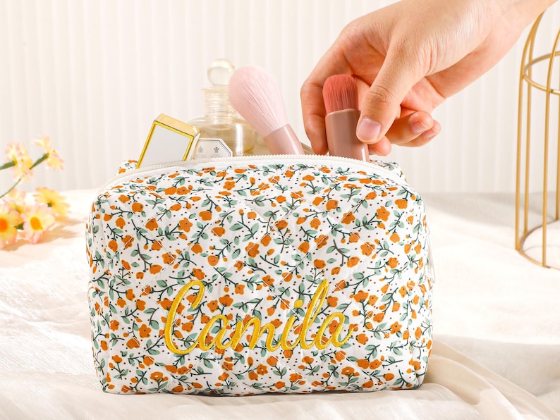 May include: A floral print cosmetic bag with the name "Camila" embroidered in yellow. The bag is filled with makeup brushes and other beauty products. The bag is white with orange and green floral details.