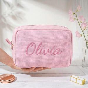 May include: A light pink rectangular cosmetic bag with a waffle-textured fabric surface. The name Olivia is embroidered in a matching pink cursive script on the front. The bag features a metal zipper closure with a matching fabric pull tab.