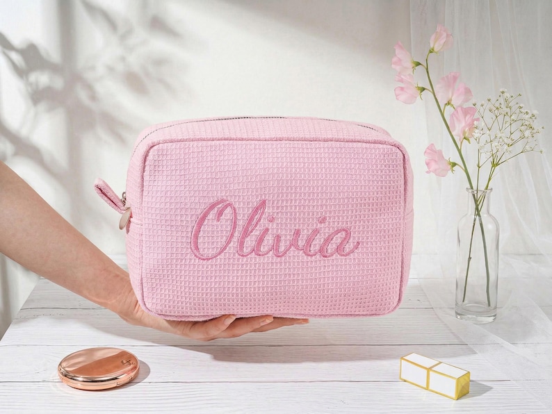 May include: A light pink rectangular cosmetic bag with a waffle-textured fabric surface. The name Olivia is embroidered in a matching pink cursive script on the front. The bag features a metal zipper closure with a matching fabric pull tab.