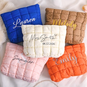 Op de afbeelding: Vijf gewatteerde make-up tasjes in blauw, bruin, wit, roze en oranje. Elk tasje heeft geborduurde tekst, waaronder namen en een datum. De tasjes liggen op een zacht, wit oppervlak.