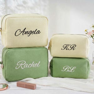 May include: Four rectangular waffle-textured toiletry bags in cream and sage green. The two larger bags feature the names Angela and Rachel embroidered in script, while the two smaller bags feature the initials KR and BL in script.