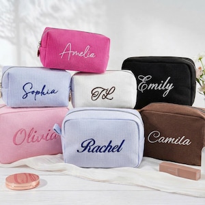 May include: A collection of rectangular cosmetic bags with a textured waffle-weave fabric. Each bag features a unique embroidered name or initials, including Amelia, Sophia, TL, Emily, Olivia, Rachel, and Camila, in various colours like pink, blue, white, and brown.