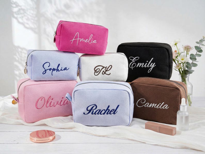 May include: A collection of rectangular cosmetic bags with a textured waffle-weave fabric. Each bag features a unique embroidered name or initials, including Amelia, Sophia, TL, Emily, Olivia, Rachel, and Camila, in various colours like pink, blue, white, and brown.