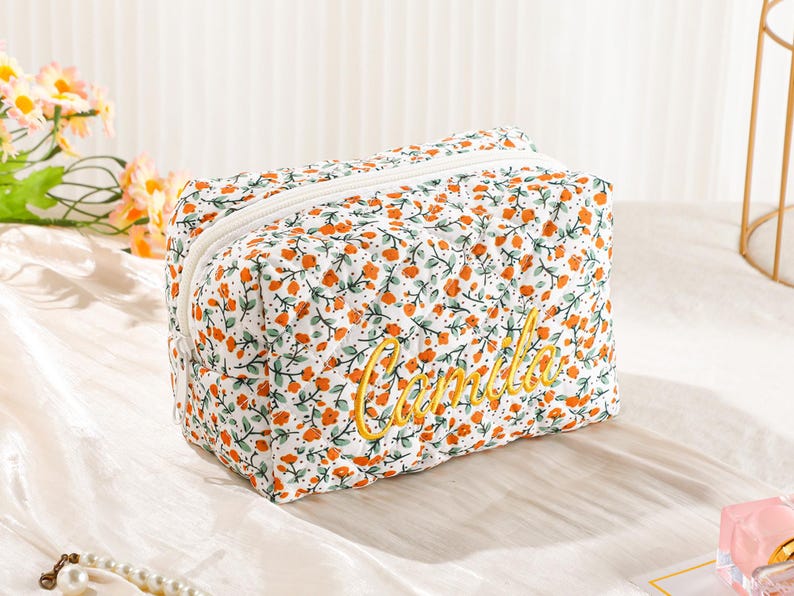 May include: A floral print cosmetic bag with a white zipper. The bag features a pattern of orange flowers and green leaves on a white background. The name "Camila" is embroidered in yellow script.