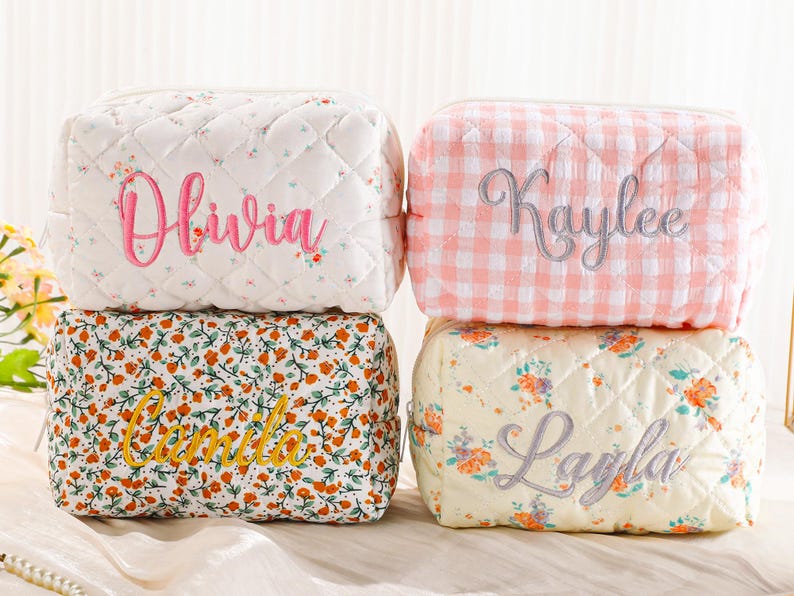 May include: Four personalized cosmetic bags in various floral and gingham patterns. The bags are quilted and embroidered with names: Olivia, Kaylee, Camila, and Layla. The bags are rectangular with a zipper closure.