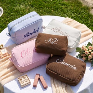 May include: Five rectangular waffle-textured cosmetic bags in light blue, white, pink, and two shades of brown. Each bag features personalised embroidery with the names Rachel, Lenita, KL, Olivia, and Camila in cursive script and silver zippers.