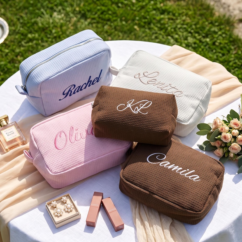 May include: Five rectangular waffle-textured cosmetic bags in light blue, white, pink, and two shades of brown. Each bag features personalised embroidery with the names Rachel, Lenita, KL, Olivia, and Camila in cursive script and silver zippers.