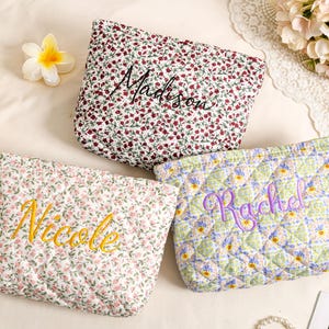 May include: Three quilted cosmetic bags with floral patterns. One bag has a red and white floral print with "Madison" embroidered in black. Another has a pink floral print with "Nicole" embroidered in yellow. The third has a green and blue floral print with "Rachel" embroidered in purple.