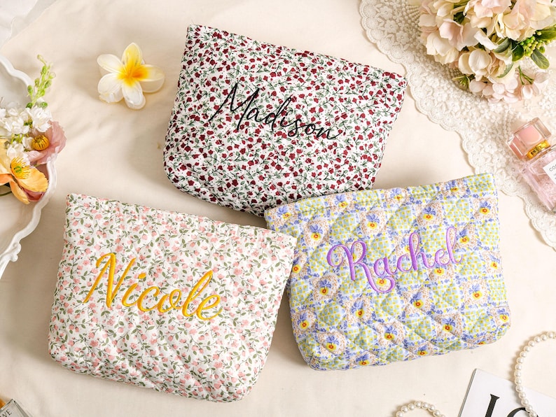 May include: Three quilted cosmetic bags with floral patterns. One bag has a red and white floral print with "Madison" embroidered in black. Another has a pink floral print with "Nicole" embroidered in yellow. The third has a green and blue floral print with "Rachel" embroidered in purple.