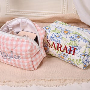 May include: Two personalized cosmetic bags. One is pink and white gingham with "Kaylee" embroidered in cursive. The other is white with blue floral print and "SARAH" embroidered in red. Both have zippers.