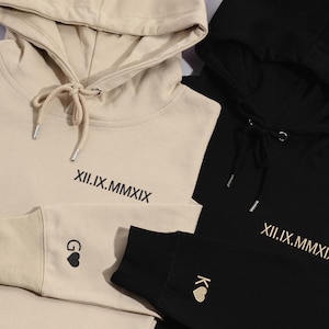 Puede incluir: Dos sudaderas con capucha, una beige y otra negra, con la fecha XII.IX.MMXIX bordada en cada una. La sudadera beige tiene un corazón negro con la letra G en la manga izquierda, y la sudadera negra tiene un corazón dorado con la letra K en la manga derecha.