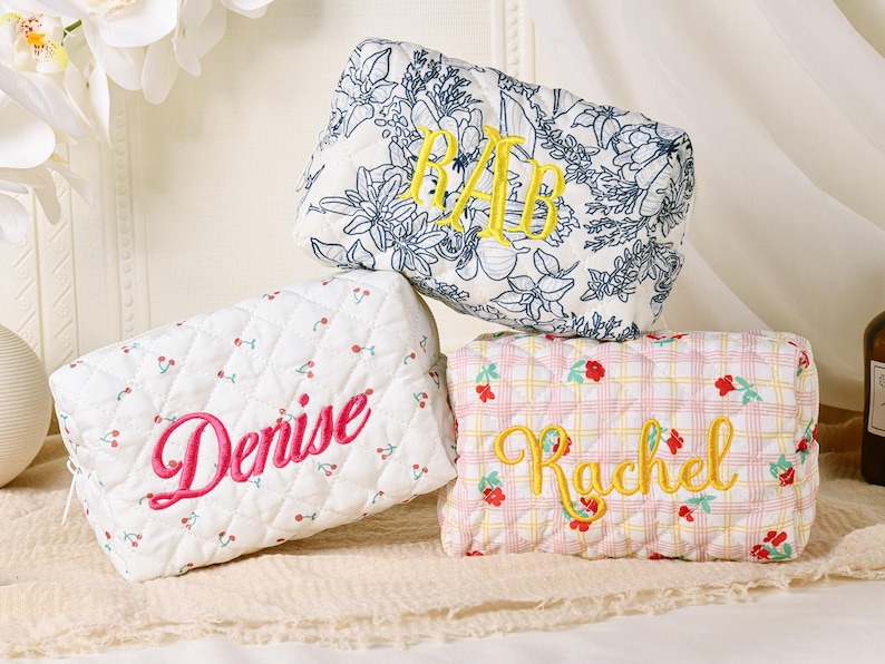 May include: Three personalized cosmetic bags. One is white with a cherry pattern and the name "Denise" embroidered in pink. Another has a floral pattern with yellow initials. The third is pink plaid with the name "Rachel" in yellow.
