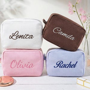 May include: Four rectangular waffle-textured cosmetic bags in white, brown, pink, and light blue. Each bag features embroidered names: Lenita in dark brown, Camila in white, Olivia in dark pink, and Rachel in dark blue, all in script font.