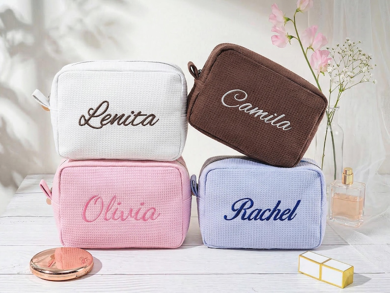 May include: Four rectangular waffle-textured cosmetic bags in white, brown, pink, and light blue. Each bag features embroidered names: Lenita in dark brown, Camila in white, Olivia in dark pink, and Rachel in dark blue, all in script font.