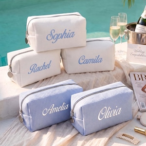 Peut inclure: Un ensemble de cinq trousses de toilette rectangulaires en tissu gaufré. Trois trousses sont blanches avec des noms brodés en bleu, et deux sont bleu clair avec des noms brodés en blanc. Chaque trousse possède une fermeture éclair argentée.