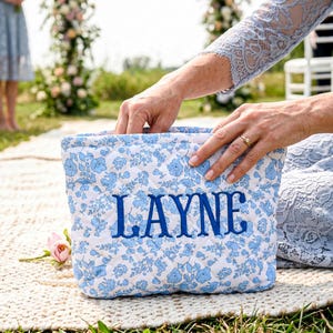 May include: A quilted, rectangular bag with a white background and a blue floral pattern. The word "LAYNE" is embroidered in blue on the front. The bag is held by a person wearing a light blue dress.