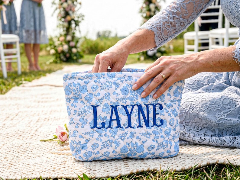 May include: A quilted, rectangular bag with a white background and a blue floral pattern. The word "LAYNE" is embroidered in blue on the front. The bag is held by a person wearing a light blue dress.