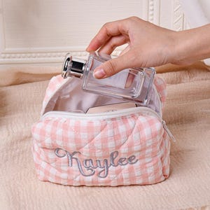 May include: A pink and white gingham cosmetic bag with the name "Kaylee" embroidered in silver thread. A clear perfume bottle with a silver cap is being placed inside the open bag. The bag is quilted and has a zipper closure.