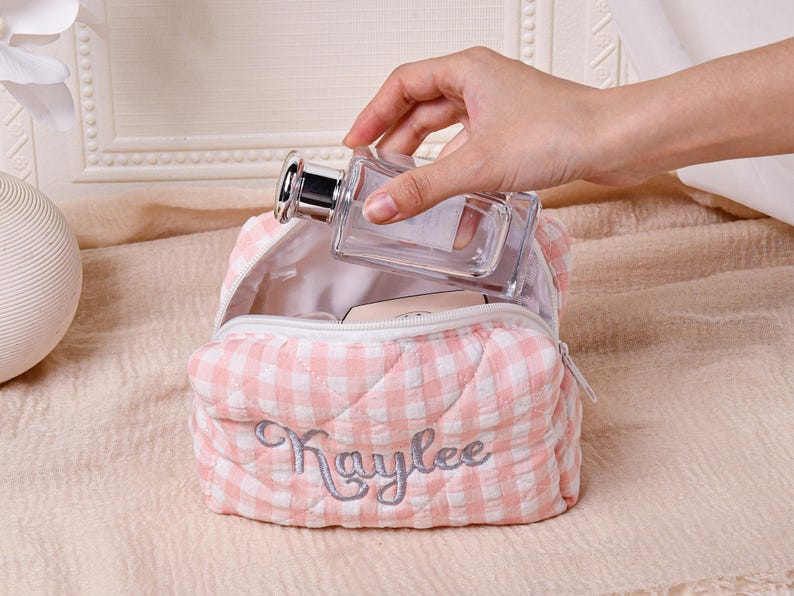 May include: A pink and white gingham cosmetic bag with the name "Kaylee" embroidered in silver thread. A clear perfume bottle with a silver cap is being placed inside the open bag. The bag is quilted and has a zipper closure.