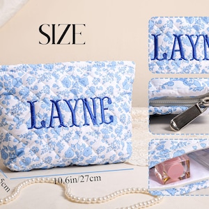May include: A quilted cosmetic bag with a blue floral pattern and the name "LAYNE" embroidered in blue. The bag is 10.6 inches long, 6.9 inches high, and 2.6 inches wide. It has a zipper closure and is designed to hold makeup and other beauty products.