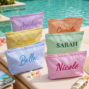 May include: A collection of colorful, striped cosmetic bags, each embroidered with a different name: Amelia, Camille, Eartha, Sarah, Bella, and Nicole. The bags are in shades of purple, orange, yellow, green, blue, and pink. The bags are on a lounge chair near a pool.