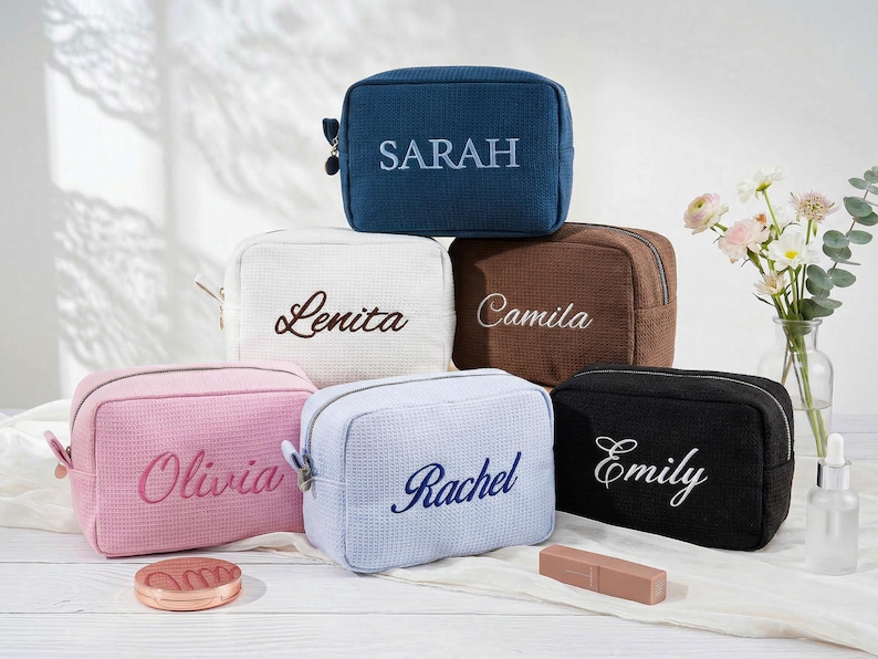 May include: A collection of six rectangular waffle-textured cosmetic bags in blue, white, brown, pink, light blue, and black. Each bag features a personalised name embroidered on the front: Sarah, Lenita, Camila, Olivia, Rachel, and Emily.