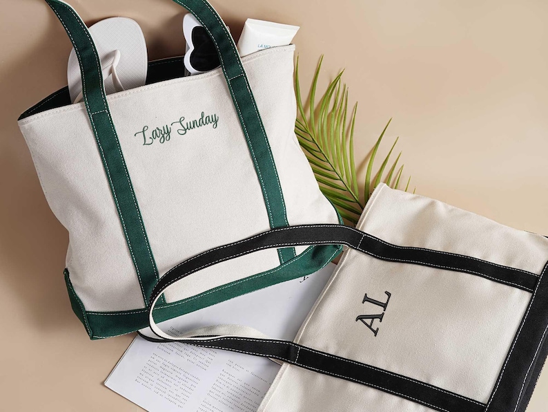 Peut inclure: Deux sacs fourre-tout en toile blanche avec des poign&eacute;es noires et vertes. Un sac a le texte "Lazy Sunday" brod&eacute; dessus, et l'autre a la lettre "AL" brod&eacute;e dessus.