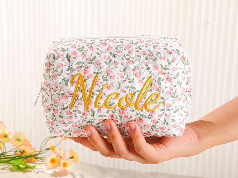May include: A floral print cosmetic bag with the name "Nicole" embroidered in gold. The bag is white with a quilted pattern and pink flower accents. It has a zipper closure and is held in a hand.