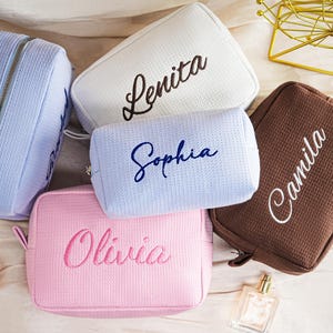 May include: A collection of personalized cosmetic bags in various colors, including light blue, white, light pink, and brown. Each bag features an embroidered name in a contrasting color: Lenita, Sophia, Olivia, and Camila. The bags have a textured waffle-like pattern.