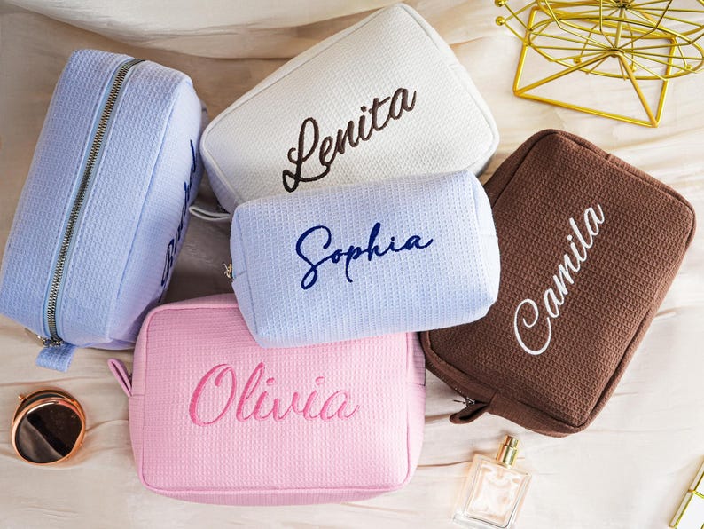 May include: A collection of five rectangular cosmetic bags with a textured waffle-knit fabric. The bags come in light blue, white, brown, and pink, each featuring a name embroidered in cursive script: Lenita, Sophia, Camila, and Olivia.