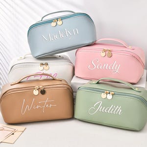 Puede incluir: Cinco neceseres personalizados en colores pastel: azul claro, blanco, rosa, beige y verde. Cada bolsa tiene una cremallera y un asa doradas, con un nombre bordado en escritura blanca: Madelyn, Sandy, Winter y Judith. Un frasco de perfume y flores están en el fondo.