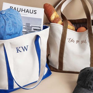 Peut inclure: Deux sacs fourre-tout en toile blanche avec des anses bleues et brunes. Un sac a les initiales "KW" brod&eacute;es sur le devant en fil bleu. L'autre sac a le texte "Let's go girls" brod&eacute; sur le devant en fil brun.