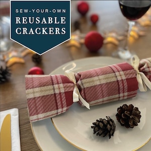 Puede incluir: Cracker de Navidad reutilizables para coser con un diseño de tela a cuadros rojo y blanco. Los crackers están atados con una cinta blanca y están sobre un plato blanco.
