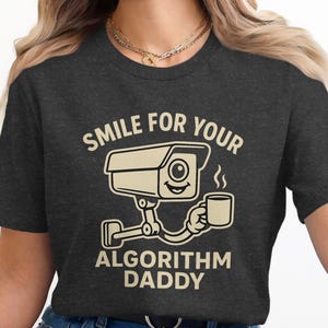 May include: Dark gray t-shirt with a humorous graphic. The design features a smiling surveillance camera holding a coffee cup, with the text "SMILE FOR YOUR ALGORITHM DADDY" in a beige color.