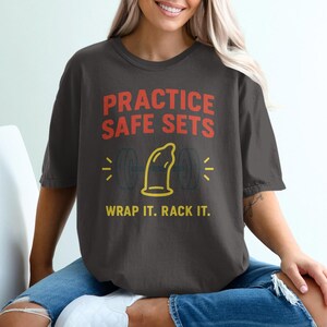 May include: Dark gray t-shirt with the text "PRACTICE SAFE SETS" in red and "WRAP IT. RACK IT." in yellow. The shirt features a yellow condom graphic with weights on either side.