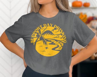 May include: Dark grey t-shirt with a yellow circular graphic that reads "SUPPORT DAY DRINKING." The graphic features a beach scene with a palm tree, water, and clouds. The shirt is a casual style.