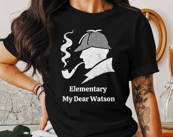 May include: Black t-shirt featuring a white graphic of a silhouette of a person with a pipe, wearing a hat, and smoking. The text "Elementary My Dear Watson" is below the graphic.