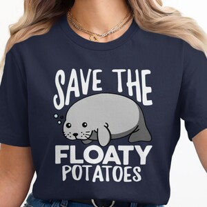 May include: A navy blue short-sleeve t-shirt featuring a graphic of a grey manatee with small bubbles. The text "SAVE THE FLOATY POTATOES" is printed in bold white capital letters above and below the manatee illustration on the front of the shirt.