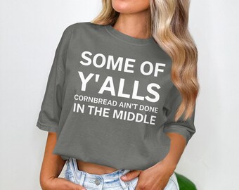 May include: Gray t-shirt with the text "SOME OF Y'ALLS CORNBREAD AIN'T DONE IN THE MIDDLE" in white block letters. The shirt has short sleeves and a crew neck. The model is wearing blue jeans.