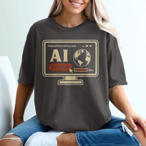 May include: Dark gray t-shirt featuring a computer graphic with the text "RebootHumanity.exe", "AI", and "REBOOT". The graphic includes a globe and a progress bar at 99%. The shirt is worn by a person.